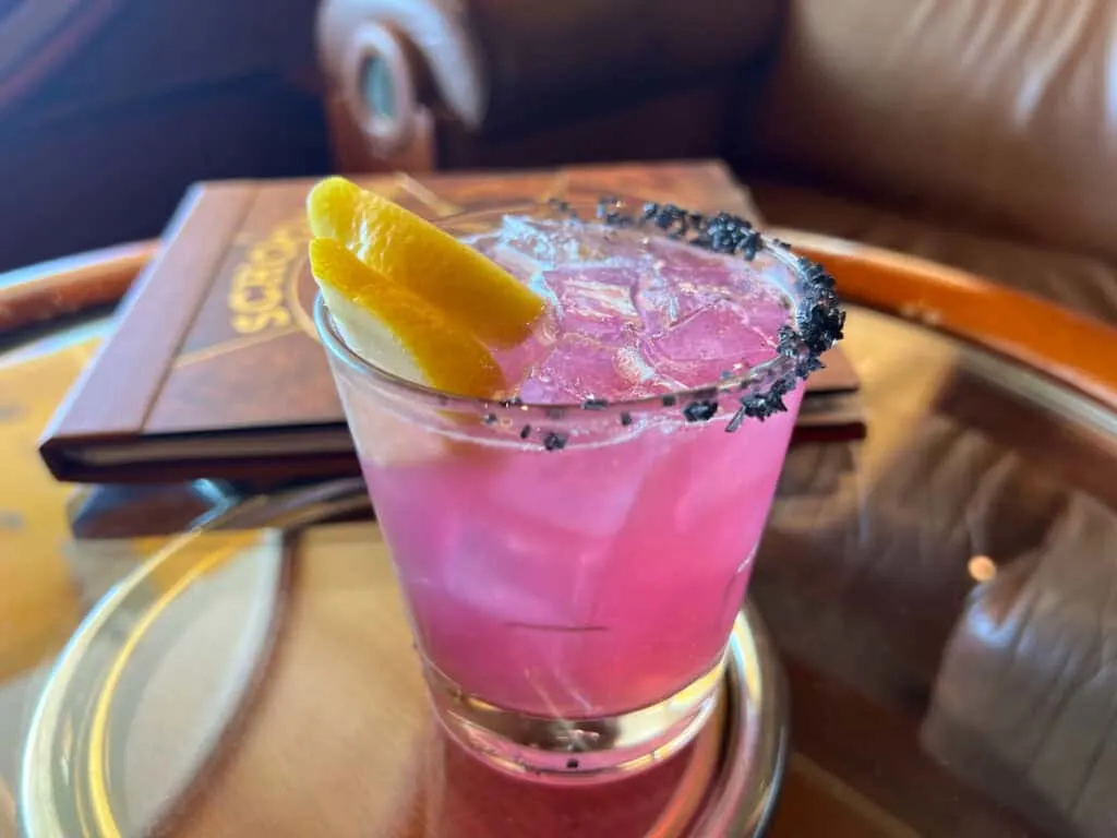 pink red liquid in glass tumbler rimmed with black salt and two lemon slices on top of round glass table with wood edge