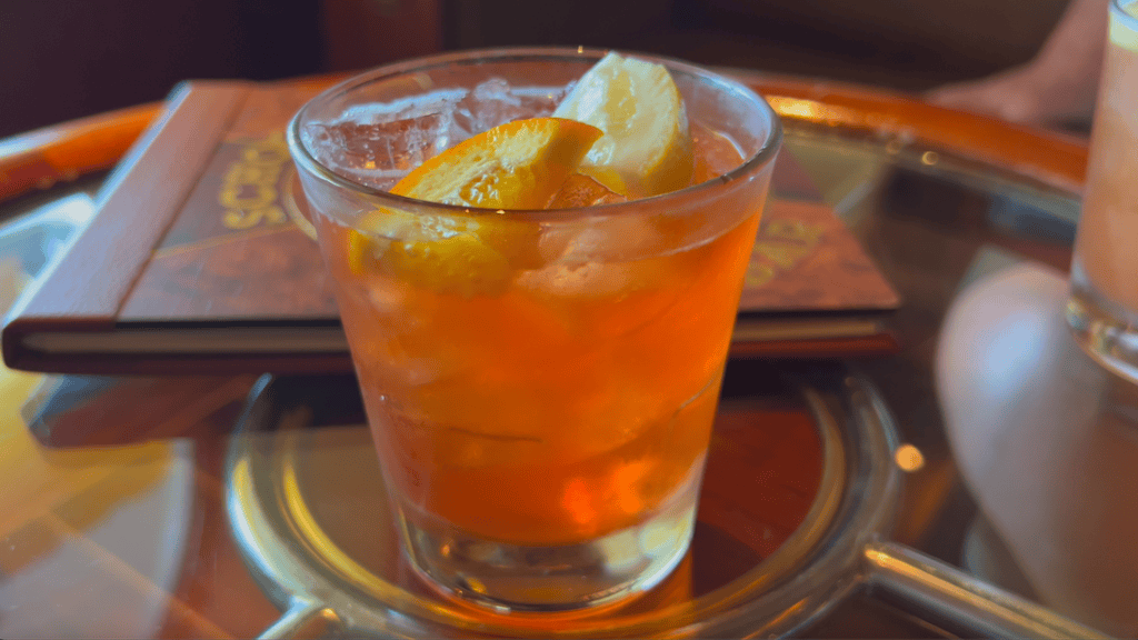 orange caramel liquid in glass tumbler with orange and lemon slice on top of round glass table with wood edge